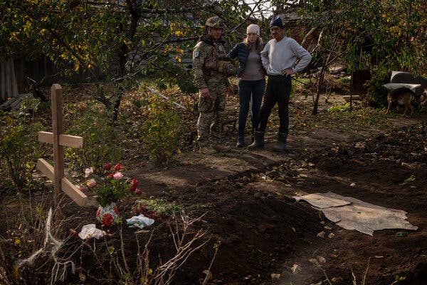 Un agente di polizia in mimetica e due civili accanto a tombe fresche contrassegnate da fiori e una croce di legno.